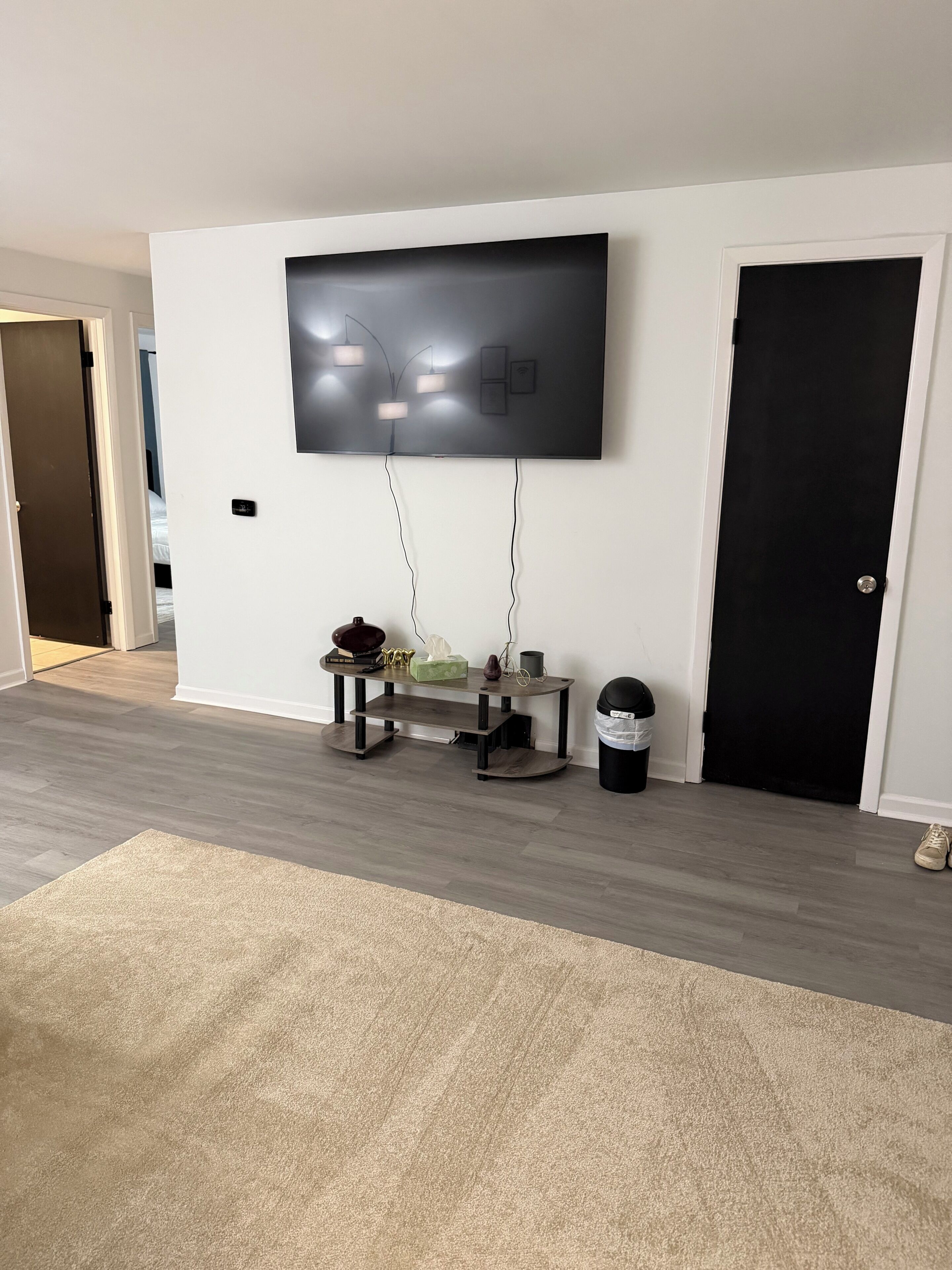 Una sala de estar con un televisor de pantalla plana montado en la pared, una mesa de centro de madera, un cesto de basura y una puerta.