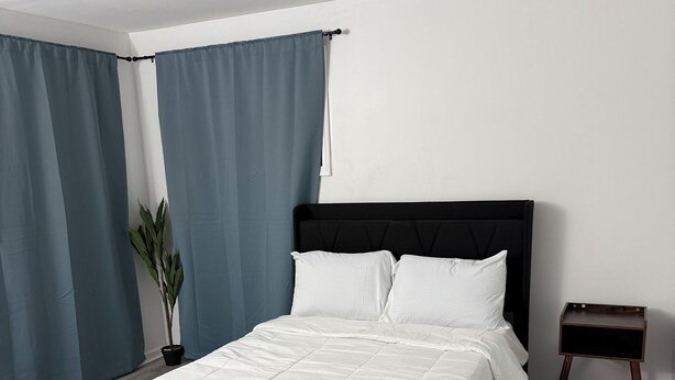 Quarto moderno com cama, cortinas, uma planta em vaso e uma pequena mesa de cabeceira.