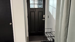 Um corredor com uma porta preta, um tapete pequeno e preto, um suporte de metal e uma cortina cinza.