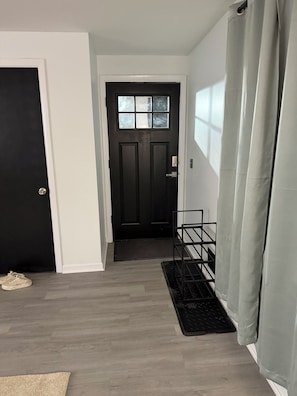 Um corredor com uma porta preta, um tapete pequeno e preto, um suporte de metal e uma cortina cinza.