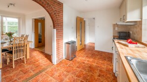 A kitchen with a brick archway, a dining area with chairs and a table, and a kitchen counter with a sink and a red pepper. - Swift Host | Watermeadow | Sleeps 5 | Parking | Garden | Dereham (488234) (Elsing)