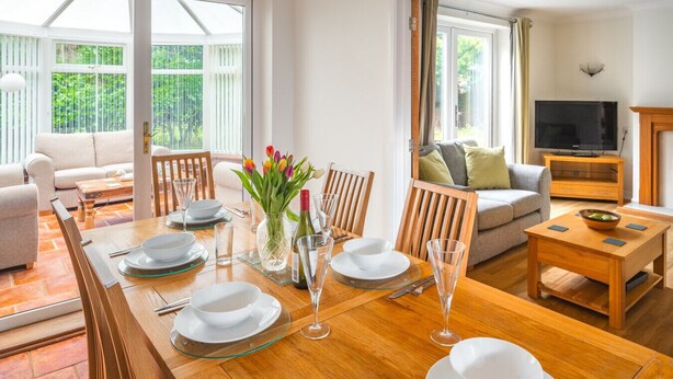 A dining area with a wooden table set for four, surrounded by chairs, a vase with flowers, and a television in the background. - Swift Host | Watermeadow | Sleeps 5 | Parking | Garden | Dereham (488234) (Elsing)
