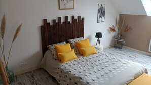 A bedroom with a wooden headboard, a bed with patterned bedding, a small side table, and a wall-mounted picture. - For a long weekend or a vacation in the countryside. (Thilouze)