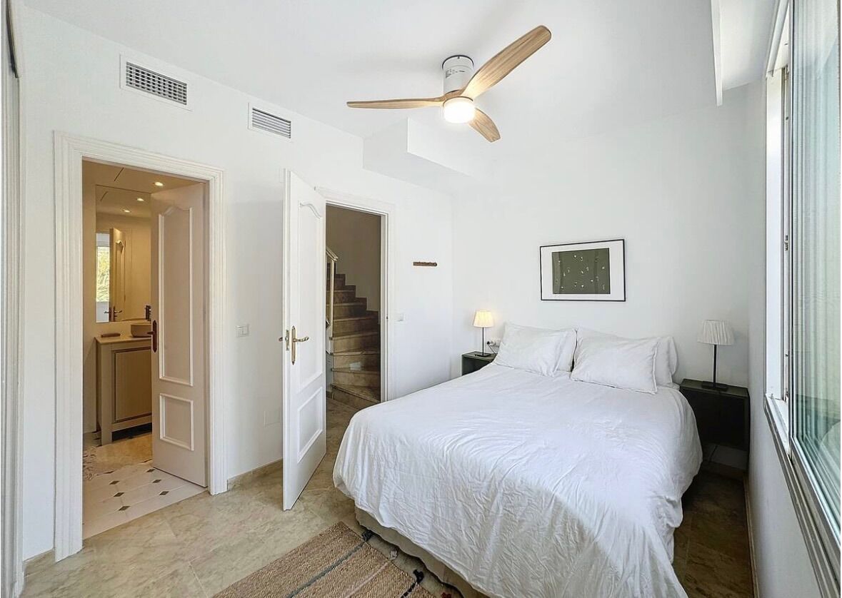 A bedroom with a bed, a ceiling fan, a nightstand, a lamp, a framed picture on the wall, and a staircase leading to another room.