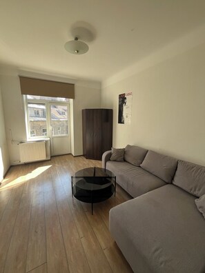 Een woonkamer met een bank, een glazen salontafel, een radiator, een raam met zonwering en een poster aan de muur.