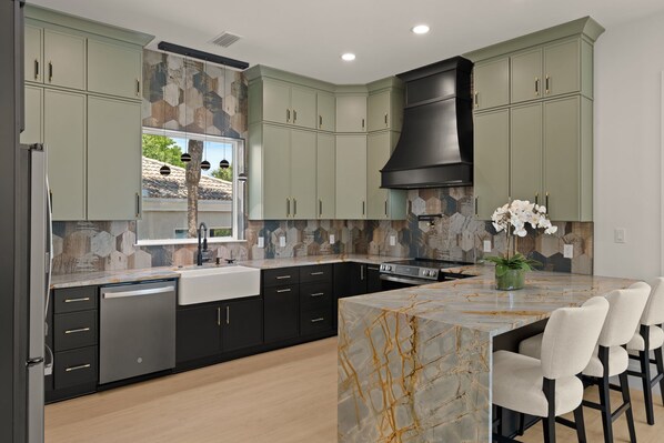 A modern kitchen with a marble island, black and green cabinets, and a built-in stove. - The Orchid Oasis: Brand New, Private Heated Pool & Beach Access (Destin)