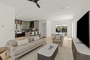 Uma sala de estar moderna com sofá, mesa de centro e televisão, integrada a uma cozinha com armários verdes e uma área de jantar.
