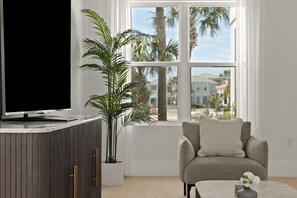 Ein modernes Wohnzimmer mit Fernseher, einer Topfpflanze, einem beigen Sofa und Blick auf Palmen und Häuser durch große Fenster.