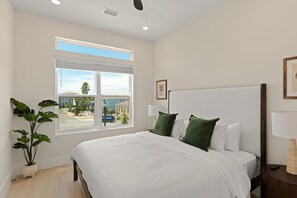 Um quarto com uma cama grande, uma janela com persianas, um vaso de planta e uma mesa de cabeceira com abajur.