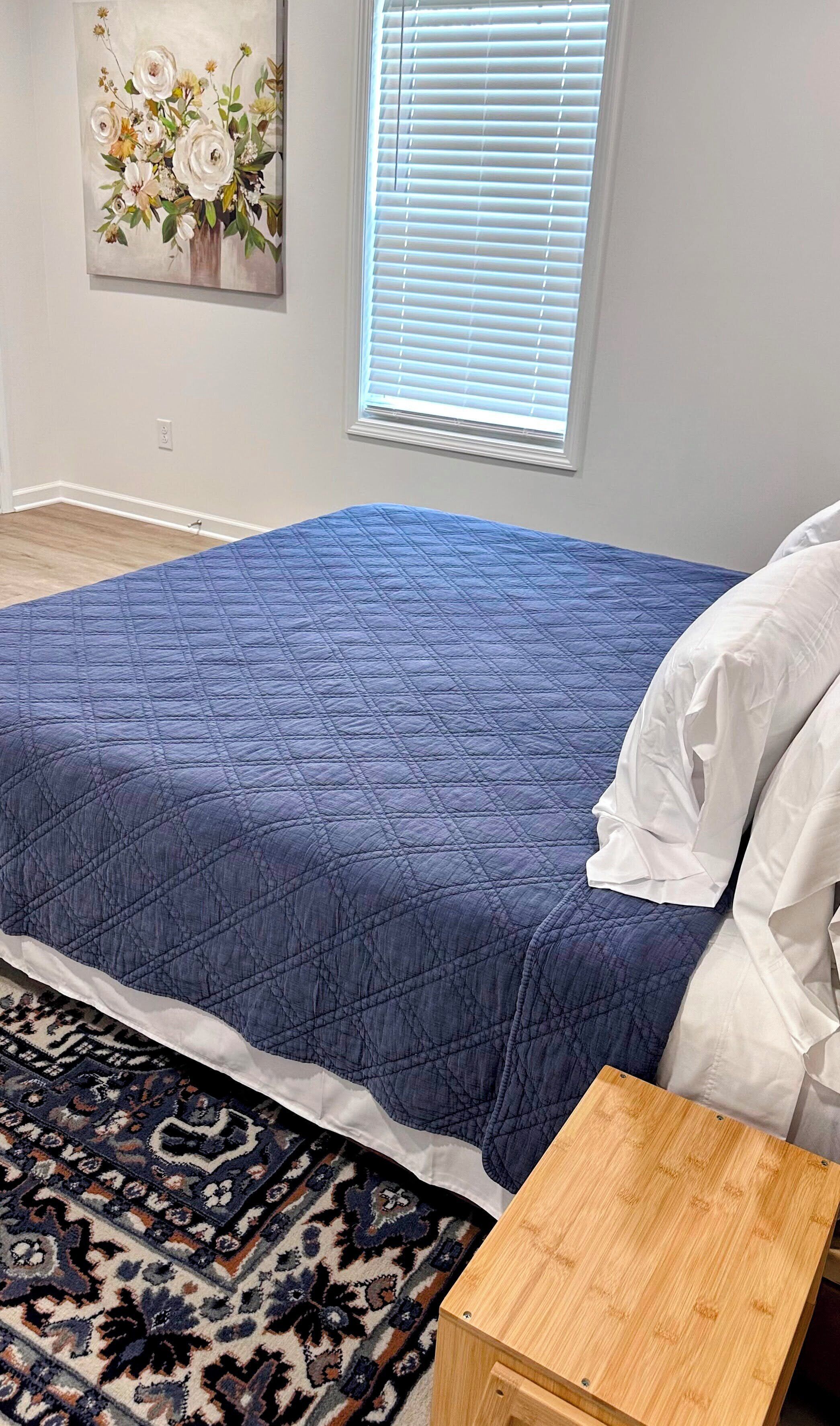 A bedroom with a blue quilted bedspread, a patterned rug, a wooden nightstand, and a wall-mounted painting.