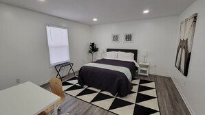 A modern bedroom with a bed, a desk, a chair, a window with blinds, a plant, and artwork on the walls.