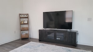 A living room with a dark wooden entertainment center, a flat-screen TV mounted above it, a woven shelf unit with decorative items, and a patterned rug on the floor.