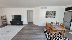 A modern living room with a flat-screen TV, a dining table with chairs, a black and white polka dot rug, and a large painting on the wall.
