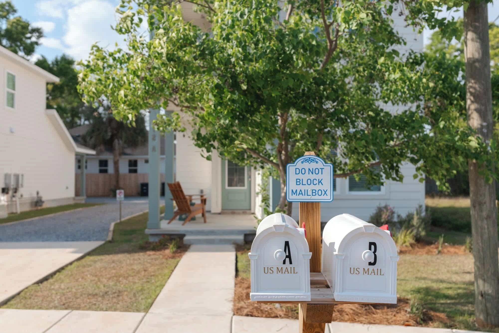 A mailbox with a 'DO NOT BLOCK MAILBOX' sign, two white mailboxes labeled 'US MAIL,' and a residential setting with a house and a tree in the background.
