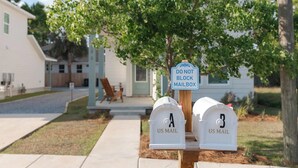 Un buzón con un letrero que dice 'NO OBSTRUIR EL BUZÓN', dos buzones blancos identificados como 'US MAIL', y un entorno residencial con una casa y un árbol al fondo.