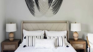 Una cama con cabecera color beige, ropa de cama blanca y almohadas estampadas en blanco y negro. Hay dos mesitas de noche con lámparas y un colgador de macramé en la pared sobre la cama.