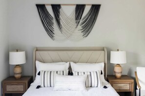 Ein Bett mit beige farbenem Kopfteil, weißer Bettwäsche und Kissen mit Schwarz-Weiß-Muster. Zwei Nachttische mit Lampen und ein Wandbehang aus Makramee befinden sich über dem Bett.