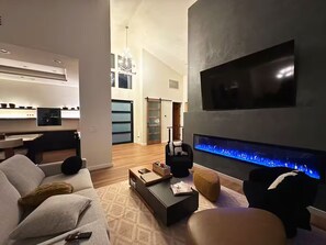 Ein modernes Wohnzimmer mit einem Flachbildfernseher, einem Kamin mit blauer Beleuchtung, einem Sofa, einem Couchtisch und einem Essbereich im Hintergrund.
