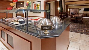 A buffet area with a sign indicating free daily breakfast, a glass display case, and a dining area with tables and chairs. - Accessible 1BR Suite | Pool & Daily Breakfast (Oconomowoc)