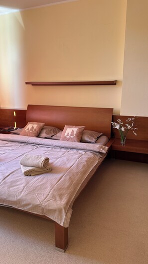 Una cama bien hecha con cabecera, una mesita de noche con un florero, y un estante en la pared sobre la cama.
