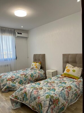 A bedroom with two beds, a nightstand, a wall-mounted air conditioner, and a window with sheer curtains. - Magnificent sea view apartment (Bir El Djir)