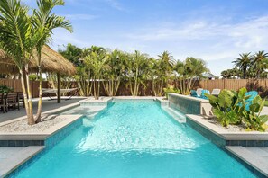 A swimming pool with a thatched roof structure, surrounded by palm trees and a wooden fence. - St. Armands Oasis | Pool, Tiki & Putting Green (Sarasota)