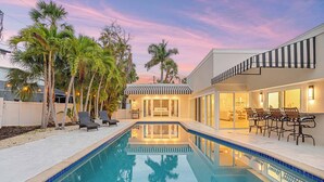 A swimming pool with lounge chairs, palm trees, and a modern house with a striped awning. - St. Armands Circle Luxe 5BR Home with Private Pool (Sarasota)
