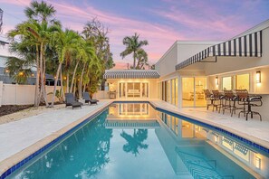 A swimming pool with lounge chairs, palm trees, and a modern house with a striped awning. - St. Armands Circle Luxe 5BR Home with Private Pool (Sarasota)