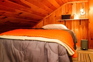 A cozy wooden room with a bed, a shelf with a lamp, and a trash bin.