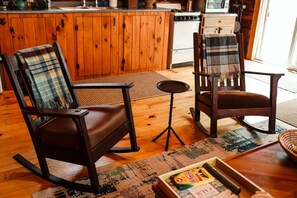 A cozy living room with wooden flooring, a rocking chair, a chair with a plaid blanket, a small round table, and a box of cereal.