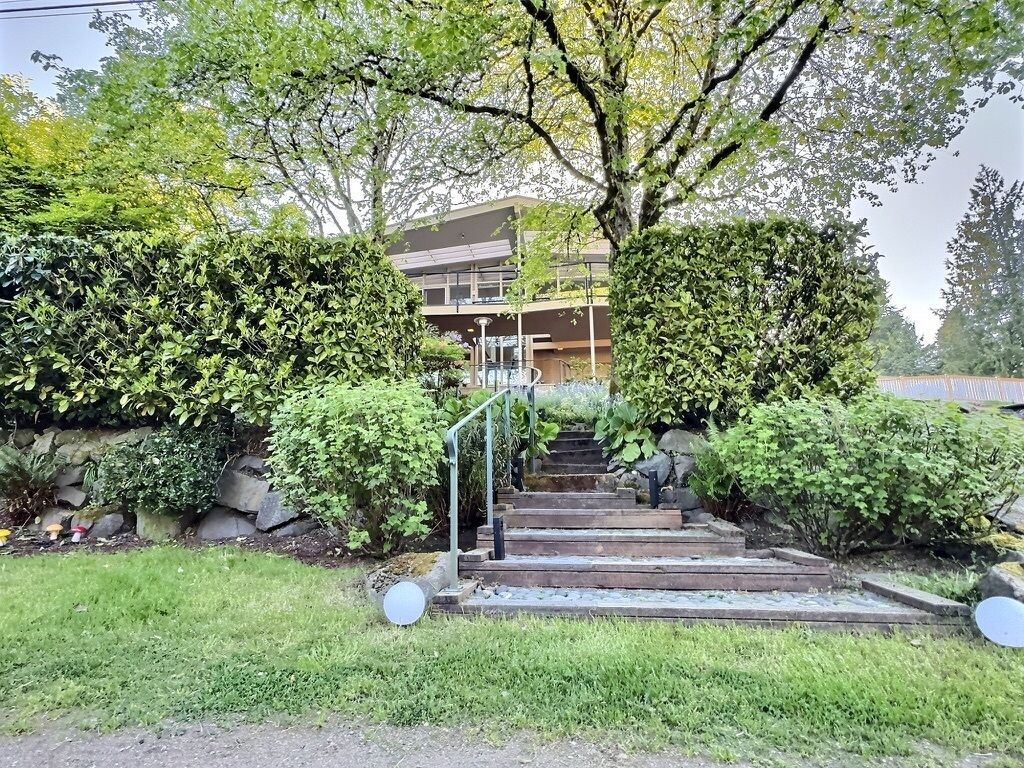 A residential building with a staircase leading to a balcony, surrounded by lush greenery and a stone wall.