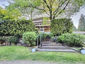 Ein Wohngebäude mit einer Treppe, die zu einem Balkon führt, umgeben von üppiger Vegetation und einer Steinmauer.