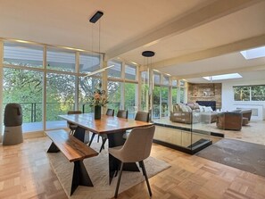 Ein moderner Essbereich mit einem Holztisch, Bänken und Stühlen, großen Fenstern und Blick ins Grüne.