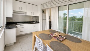 A modern kitchen with white cabinets, a black countertop, and a wooden dining table with placemats and a decorative item. - Le Cocon Bleu - 2 steps from the beach 🌴✨ (Le Moule)