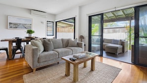 A spacious living area with a sofa, coffee table, and a dining table with chairs. There is a sliding glass door leading to an outdoor deck. - Seabrook – on Main Street, walk to beach & shops (Pottsville)