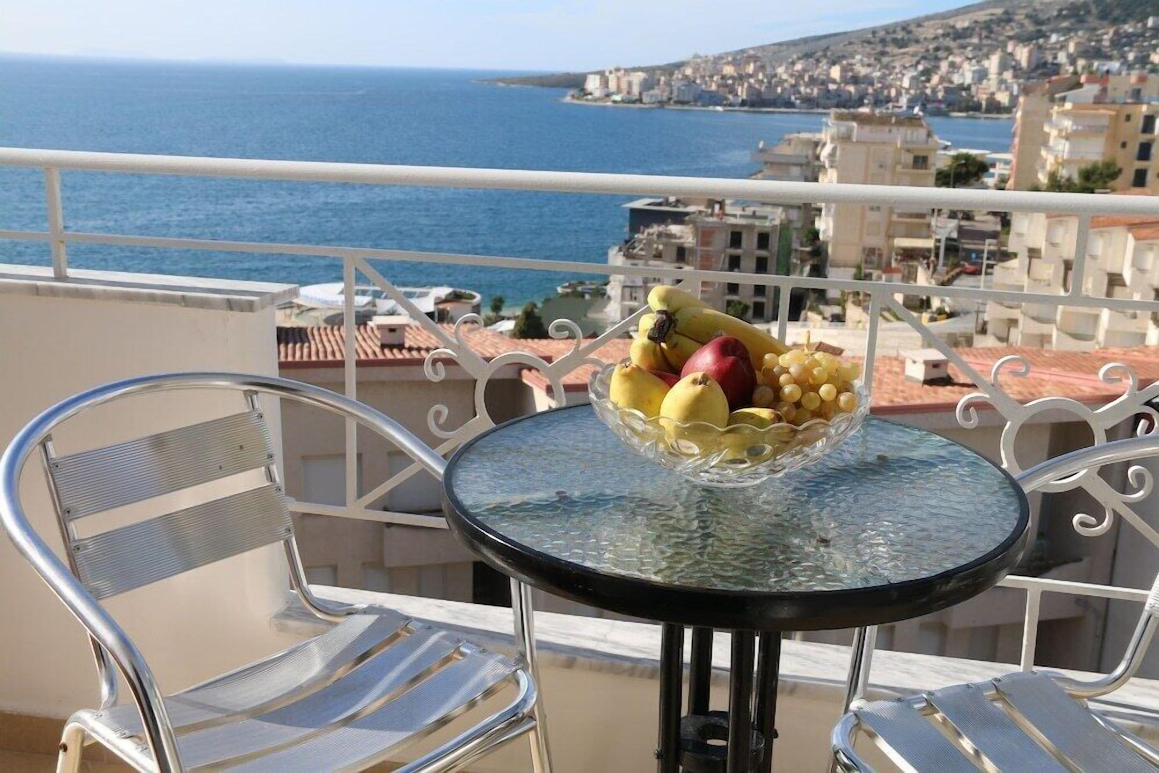 A balcony with a glass table holding fruit, metal chairs, and a view of the sea and buildings.