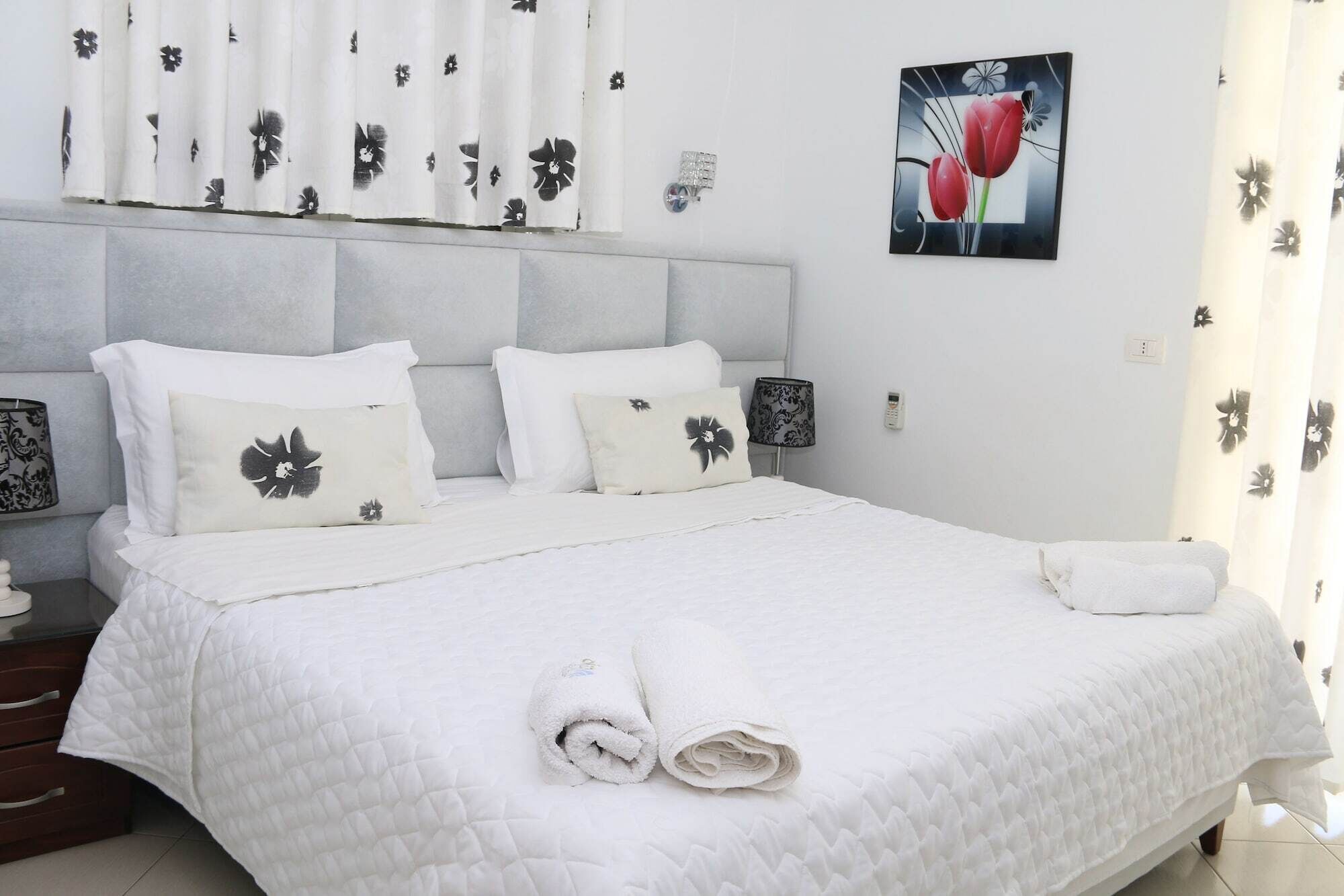 A neatly made bed with white bedding and pillows, a headboard, a wall-mounted lamp, and a framed picture of flowers.