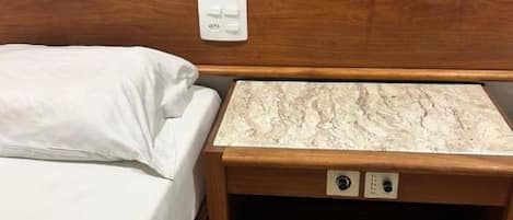 Una cama con cabecera de madera, una mesita de noche con superficie de mármol y un enchufe.
