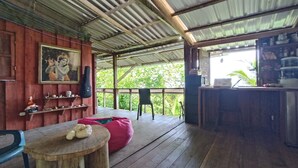 Un'area esterna riparata con un tavolo di legno, sedie, un pouf rosso, una chitarra e uno scaffale con diversi oggetti.