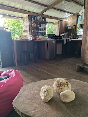 A rustic kitchen with wooden cabinets, a dining table with a red bean bag, and a wooden floor. - Casa Origen / Private oceanfront cottage (Bahía Solano)