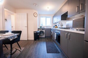 A compact kitchen with a washing machine, a dining area with a table and chairs, and a clock on the wall.