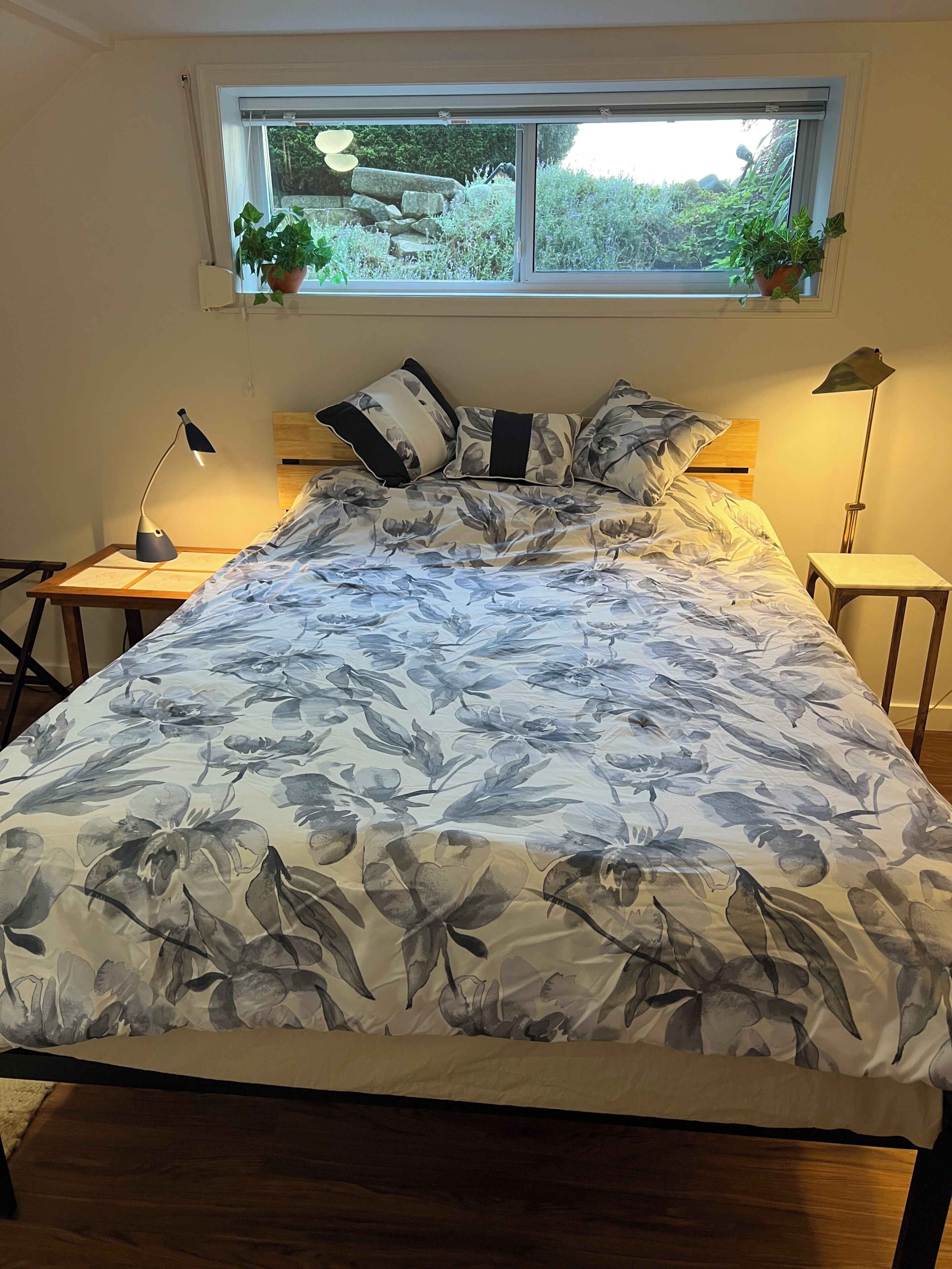 Un dormitorio con cama, ventana con plantas, mesita de noche con lámpara y velador con lámpara.
