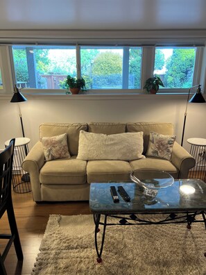 Ein Wohnzimmer mit einem beige farbenen Sofa, zwei schwarzen Stehlampen, einem Glastisch und Blick ins Grüne durch große Fenster.
