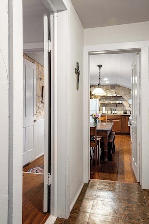 A hallway leading to a kitchen with a stone wall, wooden flooring, and a dining area with chairs and a table. - Hoerster Historic 1800s Sunday Haus, Close to Main (Fredericksburg)