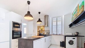 A modern kitchen with white cabinetry, a central island, a built-in oven, a microwave, pendant lights, a washing machine, and a large window. - La petite Maison Rouge, Charm and Nature in Béarn (Igon)