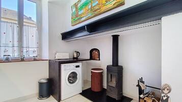 A compact kitchen with a washing machine, a wood-burning stove, and a window with curtains.