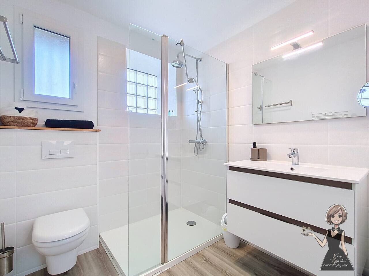 Un baño moderno con ducha envuelta en vidrio, inodoro blanco, tocador con espejo y ventana sobre el inodoro.