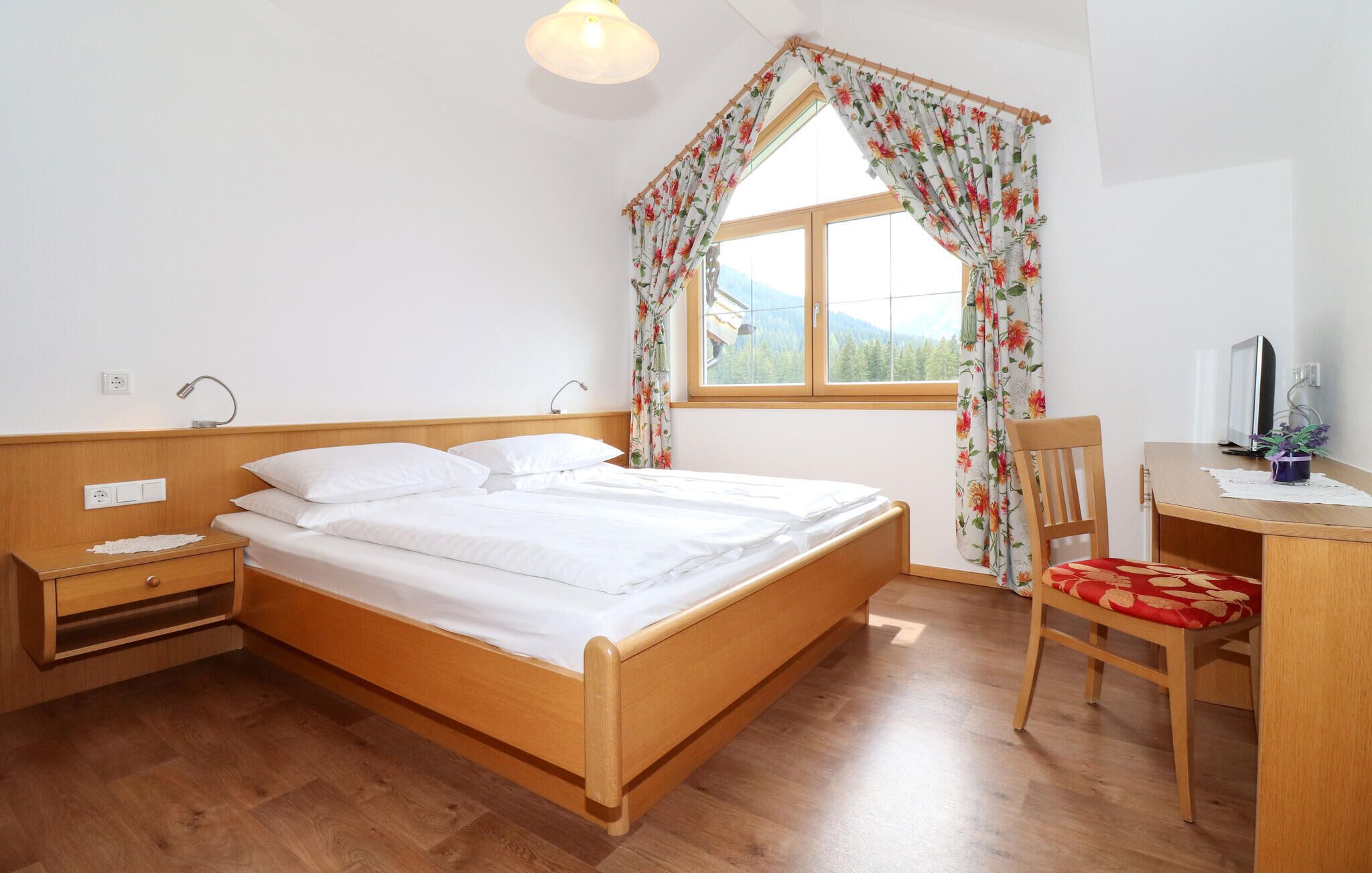 A bedroom with a wooden bed, a desk with a chair, a window with floral curtains, and a view of trees.