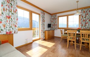 A room with wooden flooring, a dining table with chairs, a TV on a wooden cabinet, and large windows with floral curtains. - Awesome apartment in Hollersbach (Hollersbach)
