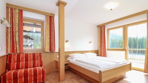 Quarto com cama, cadeira, duas janelas e vista para uma floresta.